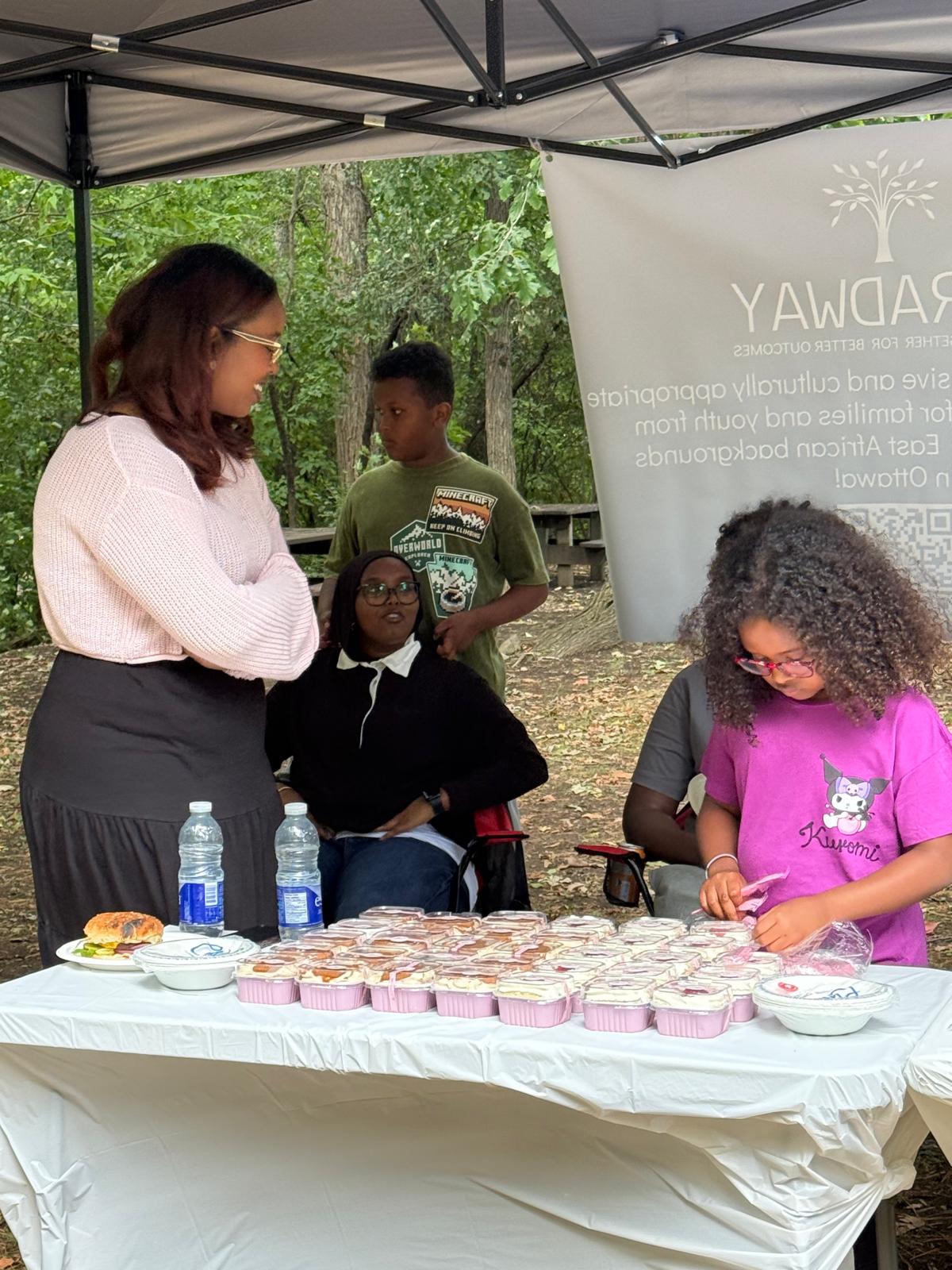 People gathered under an outdoor tent at a community event, with trays of food on a table.